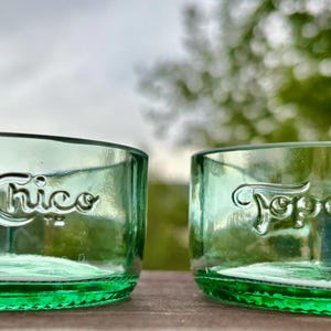 May include: Two green glass bowls, each with the embossed text "Topo Chico". The bowls are made from recycled glass bottles and have a rounded shape. The background is blurred, showing green foliage and a cloudy sky.