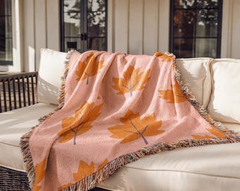 Puede incluir: Una manta tejida que presenta un color base rosa claro con un patrón repetitivo de grandes hojas de arce de color naranja dorado. La manta tiene una construcción de tela texturizada y está terminada con bordes de flecos decorativos.