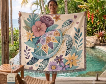 Puede incluir: Una manta tejida que presenta una tortuga marina central con un caparazón estampado, rodeada de coloridas flores tropicales y hojas en tonos rosa, amarillo y morado sobre un fondo crema, con un borde decorativo de flecos.