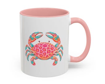 Puede incluir: Una taza de cerámica blanca con interior y asa de color rosa claro. El exterior presenta una colorida ilustración de un cangrejo con un patrón floral rosa en su caparazón, acentuado con lazos verde azulado en sus pinzas y detalles verde azulado en sus patas.
