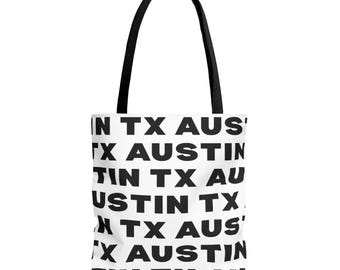 Puede incluir: Un bolso tote blanco con asas negras que presenta un patrón repetitivo del texto AUSTIN TX impreso en letras mayúsculas negras en negrita por toda la superficie del bolso. El diseño consta de varias filas con el nombre de la ciudad y el estado.