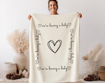 Puede incluir: Una manta de forro polar color crema con un corazón de líneas negras en el centro. El texto "we're having a baby" está impreso en letra cursiva negra a lo largo de los cuatro lados, acompañado de pequeños iconos de corazones en las esquinas.
