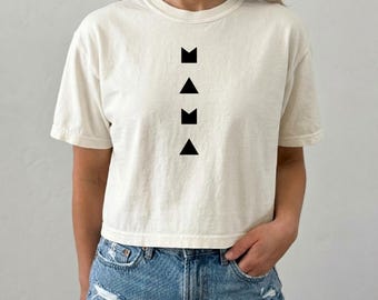Puede incluir: Una camiseta de manga corta de color crema con un diseño gráfico vertical en negro. El diseño consta de cuatro símbolos apilados que alternan entre una letra M estilizada y un triángulo, formando la palabra MAMA en un formato geométrico.