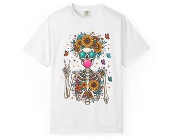 Puede incluir: Una camiseta blanca con un gráfico de un esqueleto que lleva gafas de sol de color verde azulado en forma de estrella y hace una burbuja rosa. El esqueleto está adornado con girasoles, flores naranjas y mariposas, y hace el signo de la paz.