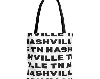 Puede incluir: Un bolso tote blanco con asas negras que presenta un patrón repetitivo del texto Nashville TN en letras mayúsculas negras y negritas. El diseño cubre toda la superficie frontal del bolso, creando un estampado tipográfico gráfico y contrastado.