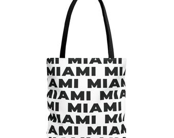 Puede incluir: Un bolso tote blanco con un patrón gráfico negro repetitivo de la palabra MIAMI impresa en una fuente sans-serif en negrita. El bolso incluye dos correas de hombro de tela negra unidas a la parte superior, creando un contraste con el blanco.