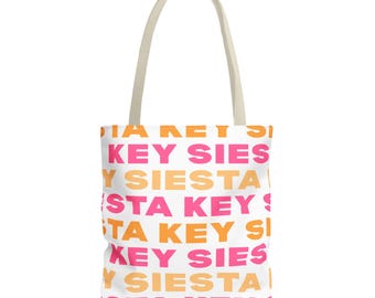 Puede incluir: Un bolso tote blanco con dos asas de hombro en color blanco roto. El bolso presenta un patrón repetitivo del texto "SIESTA KEY" impreso en filas alternas de letras mayúsculas de color rosa brillante y naranja en toda la parte frontal.