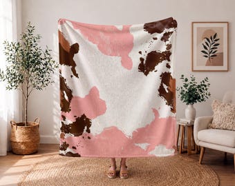 Puede incluir: Una manta suave con un patrón abstracto de manchas de vaca en tonos blanco, rosa claro y marrón oscuro. El diseño consiste en parches de color irregulares con formas orgánicas distribuidos por la superficie de la tela en un motivo repetitivo.