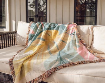 Puede incluir: Una manta tejida con un estampado botánico a gran escala de hojas de monstera en tonos amarillo, verde azulado y rosa suave. La manta rectangular tiene una construcción de tela texturizada y está terminada con un borde decorativo de flecos.