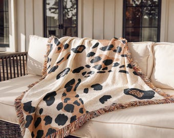 Puede incluir: Una manta tejida con una base de color crema y un patrón repetitivo de grandes manchas de leopardo irregulares en negro y marrón. La manta tiene una superficie texturizada y está rematada con flecos deshilachados multicolores en los bordes.