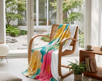 Puede incluir: Una manta suave con un vibrante estampado floral de grandes flores amarillas y rosas sobre un fondo turquesa y blanco, colocada sobre una silla de madera con respaldo de rejilla en una habitación luminosa con puertas de cristal.