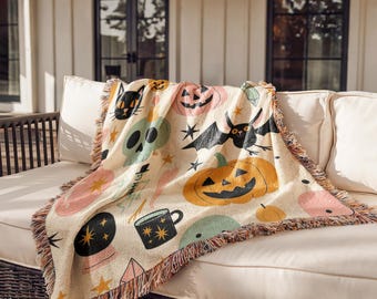 Puede incluir: Una manta tejida con fondo crema y un patrón repetitivo de ilustraciones temáticas de Halloween, que incluye calabazas naranjas, un gato negro, un murciélago volador, una calavera verde, una bola de cristal, una taza negra y pequeñas estrellas amarillas.