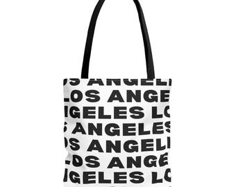 Puede incluir: Un bolso tote blanco con asas negras que presenta un patrón repetitivo del texto LOS ANGELES impreso en letras mayúsculas negras, negritas y sin remates en toda la superficie del bolso, creando un diseño gráfico de alto contraste.