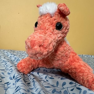 Puede incluir: Un caballo de peluche de crochet de color melocotón con pelo blanco en la cabeza y ojos negros. El caballo está tumbado sobre una colcha de cama con estampado floral azul y blanco.