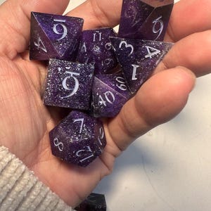 May include: A set of purple and white dice with a galaxy design. The dice are in various shapes, including d4, d6, d8, d10, d12, and d20. The numbers are white and easy to read. Ideal for tabletop games.