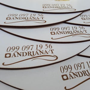 May include: A stack of white round wooden coasters with a brown border and the text "099 097 19 56 ANDRIANA T" engraved on each coaster.