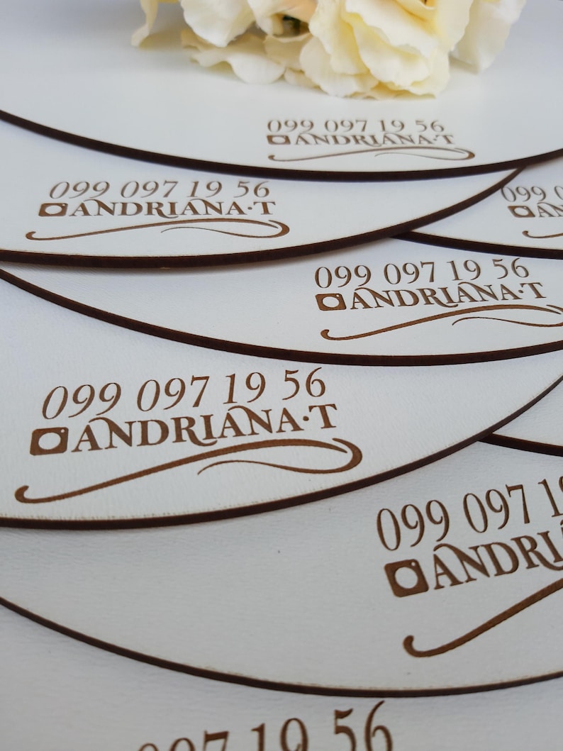 May include: A stack of white round wooden coasters with a brown border and the text "099 097 19 56 ANDRIANA T" engraved on each coaster.