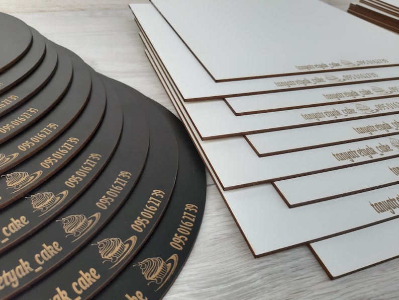 May include: A stack of black round cake boards with the text "095 016 27 39" and a cupcake graphic printed on each board. The boards are stacked on top of each other, with the largest board at the bottom and the smallest board at the top. The boards are all the same size, but they are stacked in a way that makes them look like they are getting smaller as they go up. The boards are all black, and they have a smooth, glossy finish.