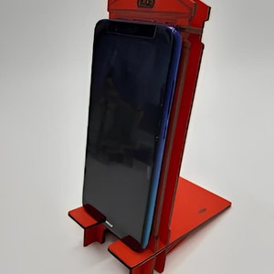 May include: Red phone booth shaped phone stand with a black smartphone in the stand.