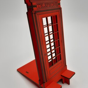 May include: Red wooden phone booth stand with a crown on top and the word "TELEPHONE" printed on the front. The stand is designed to hold a phone or other device.