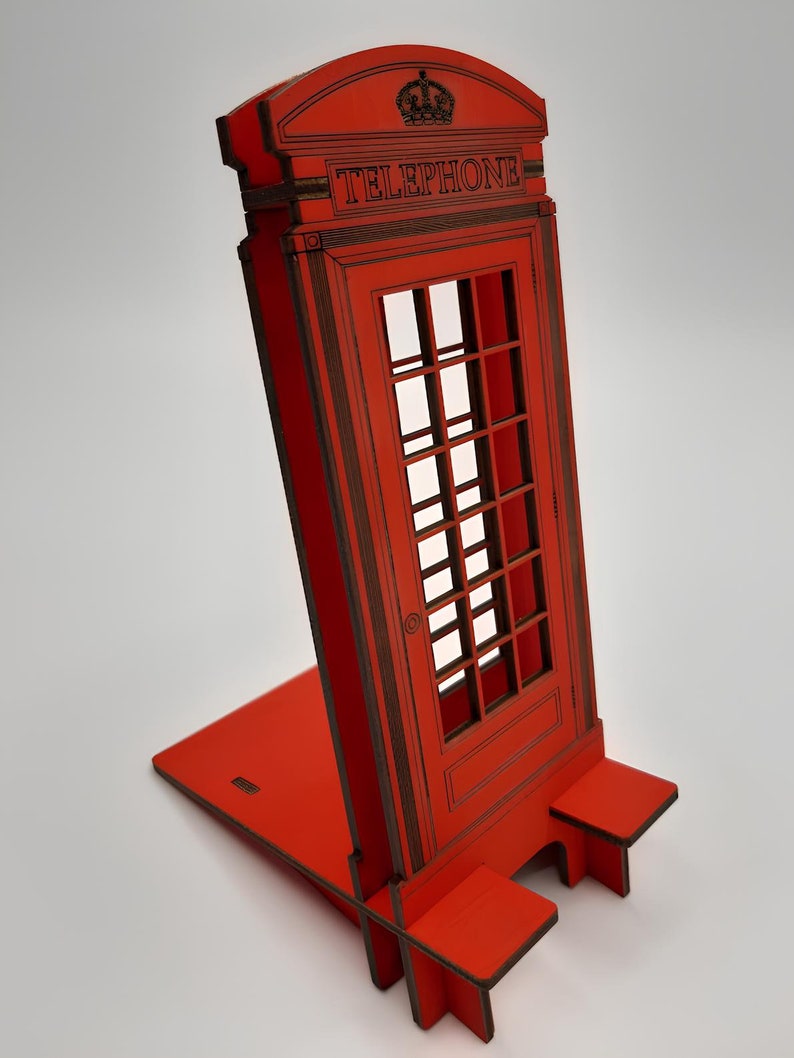 May include: Red wooden phone booth stand with a crown on top and the word "TELEPHONE" printed on the front. The stand is designed to hold a phone or other device.