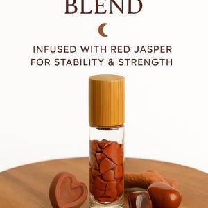 May include: A clear glass roller bottle with a bamboo cap, filled with red jasper stones. The text "GROUNDING BLEND" and "INFUSED WITH RED JASPER FOR STABILITY & STRENGTH" are displayed. Also, a heart-shaped stone and other red stones are on a wooden surface.