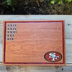 May include: A rectangular wooden serving board featuring a red resin border. The board displays the San Francisco 49ers logo in the bottom right corner and a vertical column of Roman numerals XXIX, XXIV, XXIII, XVI, and XIX on the left side.