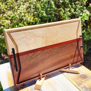 May include: A wooden serving tray with a light wood top and a dark wood bottom. The tray has two black metal handles and a stand made of light wood.
