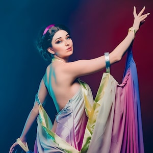 Puede incluir: Una mujer con un vestido fluido y colorido con una diadema rosa. El vestido está hecho de una tela transparente y tiene un degradado de colores, incluyendo azul, morado y amarillo. La mujer está posando con el brazo levantado, y la tela del vestido fluye a su alrededor.