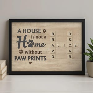 Könnte beinhalten: Gerahmtes Holzschild mit dem Text "A HOUSE is not a Home without PAW PRINTS." Das Wort "Home" ist mit einem Pfotenabdruck stilisiert. Scrabble-Kacheln buchstabieren Namen. Der Rahmen ist schwarz, und der Hintergrund ist eine helle Holzmaserung.