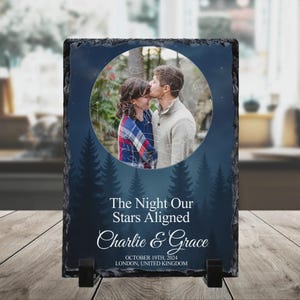 Könnte beinhalten: Rechteckige Schiefertafel mit einem Foto eines sich küssenden Paares, vor einem Sternenhimmel und einer Waldsilhouette. Der Text "The Night Our Stars Aligned" und die Namen "Charlie & Grace" sind zusammen mit Datum und Ort abgebildet.