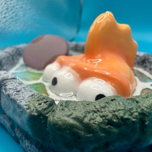 May include: A ceramic figurine of a cartoon fish with large eyes and an orange body. The fish is partially submerged in a green and blue ceramic pond.