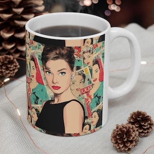 May include: White ceramic coffee mug featuring a pop art style image of Audrey Hepburn in a black dress. The mug's design incorporates a collage of vintage magazine covers and portraits, creating a retro aesthetic. The mug is filled with a dark liquid.