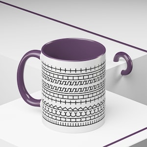 May include: White ceramic mug with a purple interior and handle. The exterior features a repeating black geometric pattern. The mug is displayed on a white surface with a purple accent.