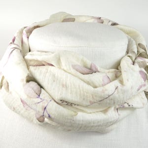 Könnte beinhalten: Cremefarbener Infinity-Schal mit floralem Muster. Der Schal hat ein sich wiederholendes Design aus lila und grünen Blättern und Stielen. Der Stoff ist um einen weißen Mannequinkopf drapiert und zeigt die Form und Textur des Schals.