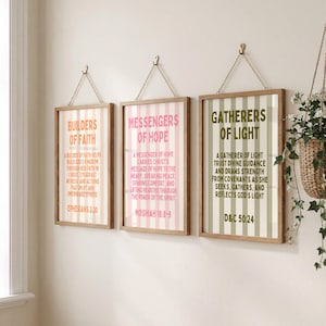 Puede incluir: Tres impresiones enmarcadas con texto inspirador cuelgan de una pared. Cada impresión tiene un marco de madera y está suspendida por una cadena. Las impresiones presentan rayas verticales en rosa, verde y melocotón, con texto correspondiente.