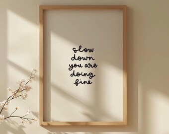 May include: A framed print with the words "slow down you are doing fine" in black script. The frame is light brown, and the background is a neutral beige. A sprig of white flowers is in the lower left corner.