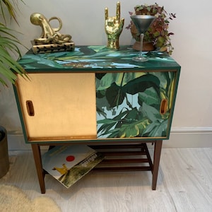 May include: A vintage wooden cabinet with a sliding door. The cabinet is painted green and has a tropical leaf pattern. The cabinet has a shelf and is decorated with gold figurines.