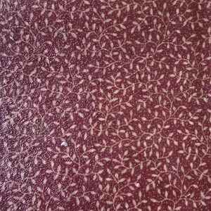 May include: Close-up of a burgundy fabric with a repeating pattern of delicate, white, leaf-like designs. The fabric has a subtle, textured appearance, with a slight sheen. The overall effect is one of understated elegance and natural motifs.