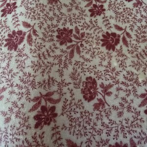 May include: Close-up of a fabric with a repeating floral pattern. The design features burgundy flowers and foliage on a cream-colored background. The fabric has a soft, textured appearance, suggesting a textile like velvet or a similar material.
