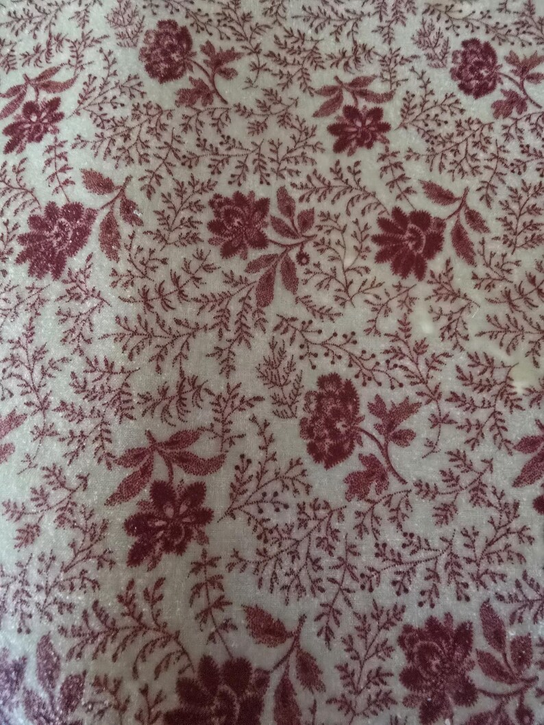 May include: Close-up of a fabric with a repeating floral pattern. The design features burgundy flowers and foliage on a cream-colored background. The fabric has a soft, textured appearance, suggesting a textile like velvet or a similar material.