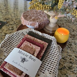 May include: A stack of Moxie Bee reusable beeswax wraps in various floral patterns and colors, including red and brown. The wraps are bundled together with a white label featuring a bee illustration. The wraps are on a wooden tray with a crocheted doily.