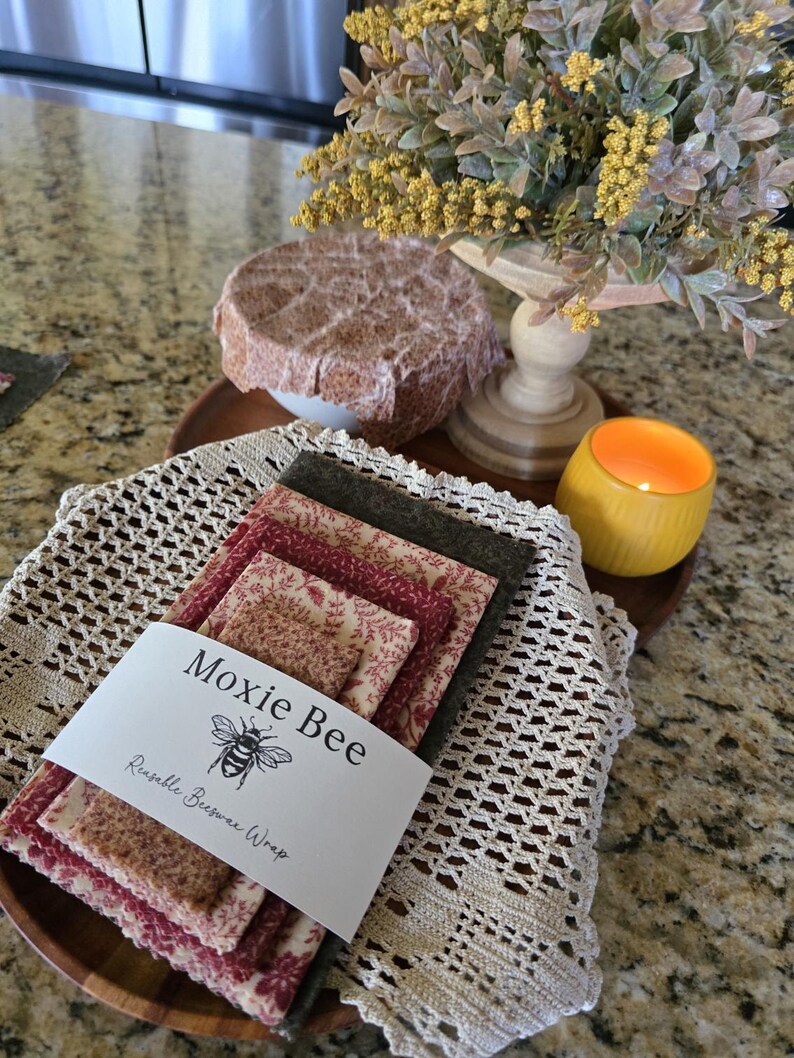 May include: A stack of Moxie Bee reusable beeswax wraps in various floral patterns and colors, including red and brown. The wraps are bundled together with a white label featuring a bee illustration. The wraps are on a wooden tray with a crocheted doily.