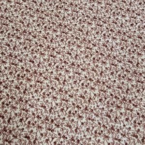 May include: Close-up of a fabric with a repeating pattern of small, dark red floral sprigs on a cream-colored background. The design features delicate leaves and tiny berries, creating a dense, all-over print. The fabric appears to be cotton or a similar material.