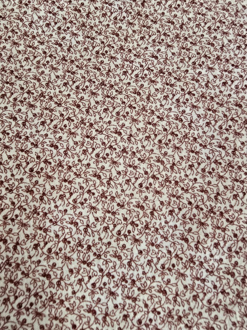 May include: Close-up of a fabric with a repeating pattern of small, dark red floral sprigs on a cream-colored background. The design features delicate leaves and tiny berries, creating a dense, all-over print. The fabric appears to be cotton or a similar material.