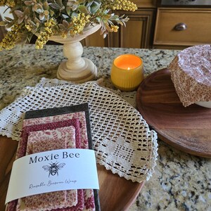 May include: A collection of reusable beeswax wraps with a floral pattern, stacked on a wooden tray. The wraps are labeled "Moxie Bee" with a bee illustration. A lit yellow candle and a bowl covered with a patterned wrap are also visible.