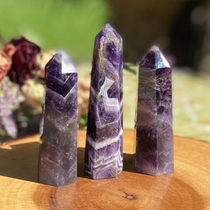 May include: Three amethyst crystal towers with varying heights and shapes. The crystals display a mix of purple and white bands, with a polished, smooth surface. The crystals are standing on a wooden surface, with a blurred background of greenery and dried flowers.