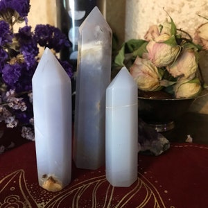May include: Three light blue crystal towers of varying sizes. The crystals have a smooth, polished surface and are arranged in a vertical composition. Dried purple flowers and a bowl of dried roses are in the background.