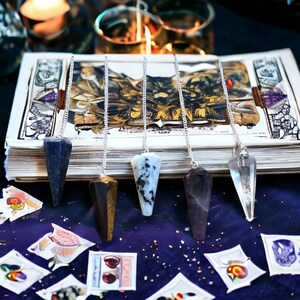 May include: Five silver chain pendulums with different colored crystal points. The pendulums are arranged in a row on a book with a dark blue background. The crystals are black, brown, white, clear, and dark blue.