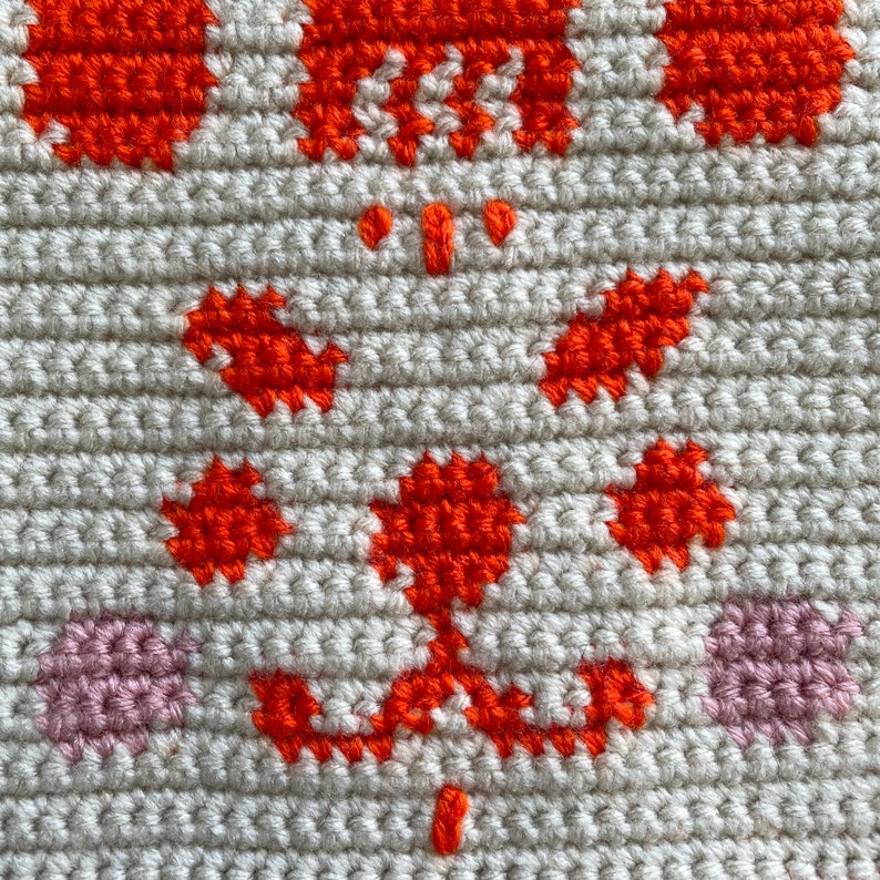 K&ouml;nnte beinhalten: Nahaufnahme eines geh&auml;kelten Stoffes mit einem Muster aus orangefarbenen und rosafarbenen Blumen. Der Stoff ist hellbeige.